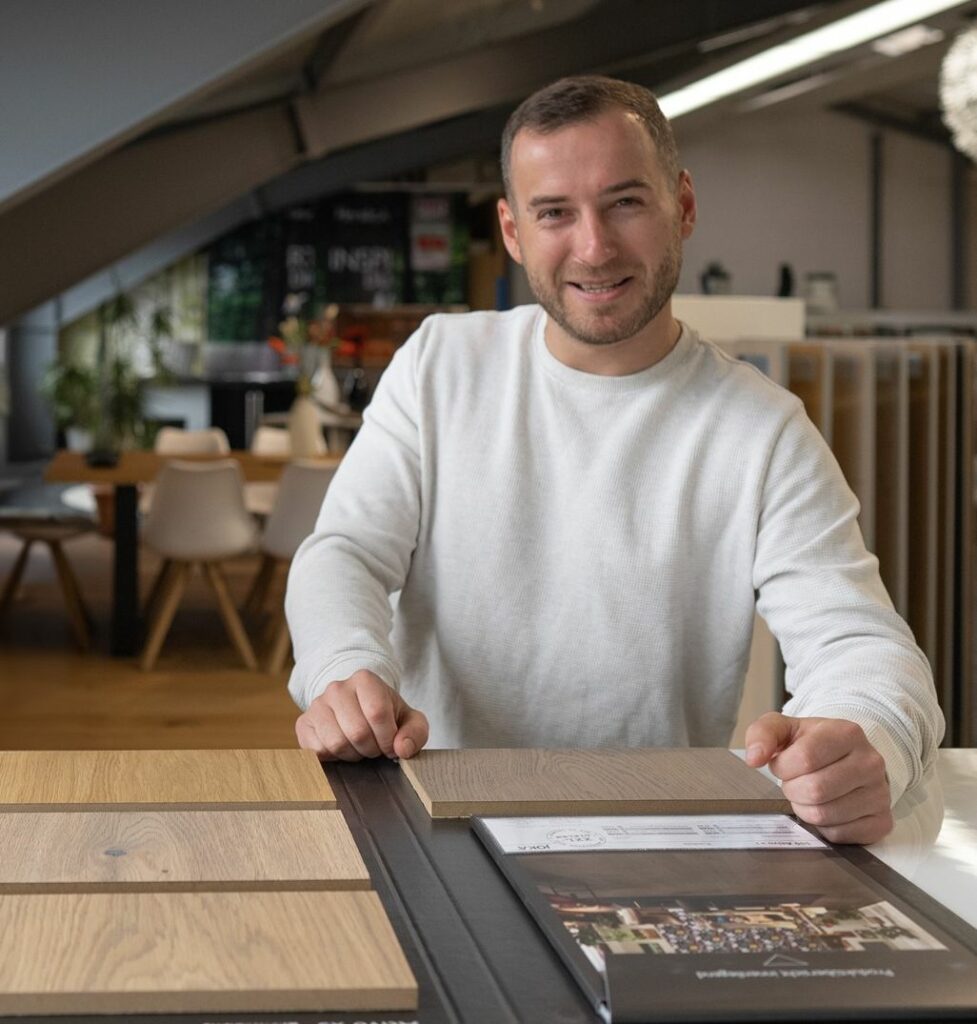 Ein Mann in einem weißen Pullover sitzt an einem Tisch in einem modernen Ausstellungsraum, an dem Holzbodenmuster und eine Broschüre ausgestellt sind. - Firmenadresse