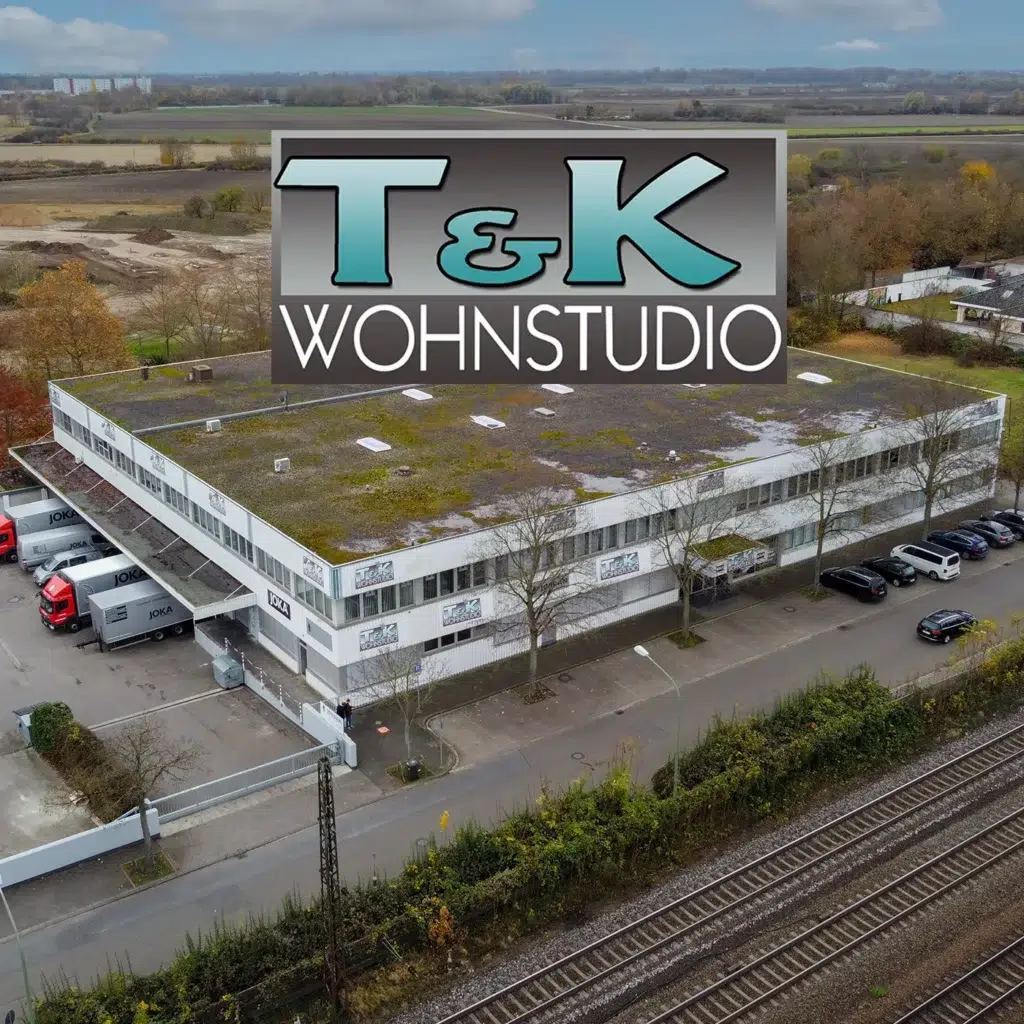 Aus der Luft ist ein rechteckiges Gebäude mit einem markanten „T&K Wohnstudio“-Schild auf dem Dach zu erkennen. In der Nähe parken mehrere Fahrzeuge, eingebettet zwischen Bahngleisen und Feldern, was eine Atmosphäre hervorruft, die der Mischung aus Zweckmäßigkeit und malerischer Umgebung von Schads Wohnträume ähnelt. - SCHADS Wohnträume - Raumausstattung, Bodenbeläge, Vinylboden, Parkett, Gardinen - Josef-Beyerle-Straße 11, 71263 Weil der Stadt