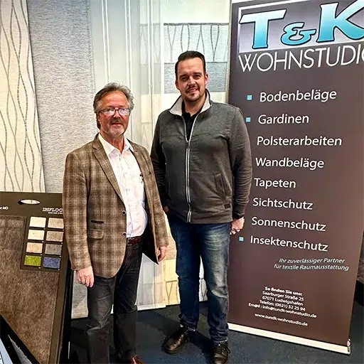 Zwei Männer stehen neben einem Display mit einem Banner von T&K Wohnstudio und präsentieren stolz ihr Sortiment an Bodenbelägen und Jalousien. Entdecken Sie mehr von Über Uns, wo sie die Handwerkskunst hinter ihren berühmten Designs präsentieren. - Firmenadresse