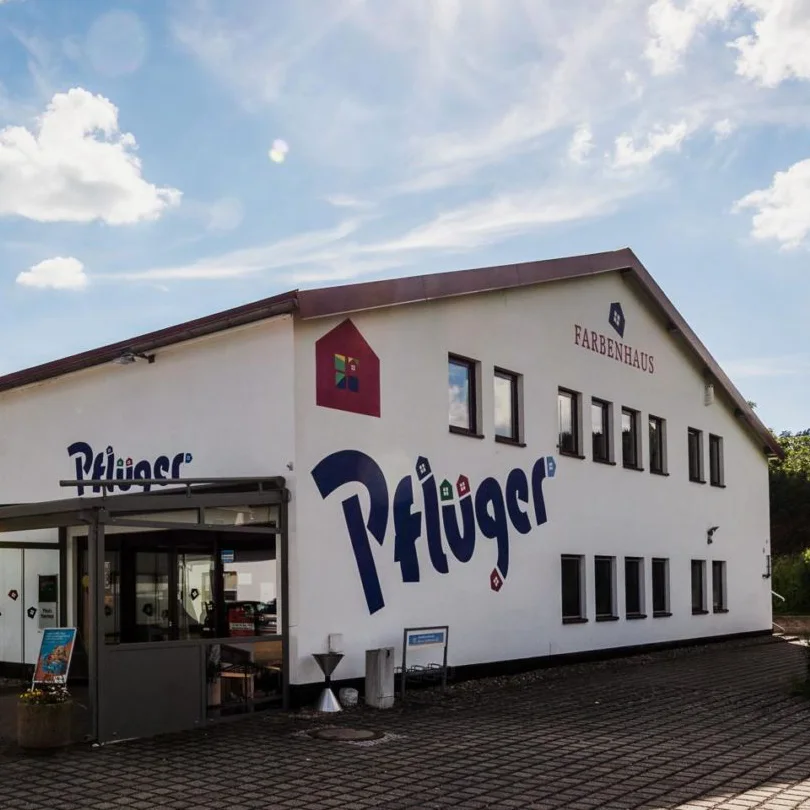 Ein weißes zweistöckiges Gebäude mit den Aufschriften „Pflüger“, „FARBENHAUS“ und „Schads Wohnträume“ an der Außenseite, mit vielen Fenstern und einer gepflasterten Fläche davor unter einem leicht bewölkten Himmel. - Farbenhaus Pflüger - Raumausstattung, Bodenbeläge, Vinylboden, Parkett, Farben, Malerarbeiten - Simmenet 2, 37235 Hessisch Lichtenau