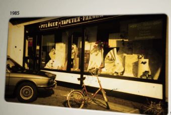 Ein Fahrrad steht auf dem Bürgersteig vor einem Geschäft mit großen Schaufenstern, in denen verschiedene Artikel ausgestellt sind. Links ist ein Auto teilweise zu sehen. Das Foto mit der Überschrift „1985“ zeigt eine klassische Straßenszene – perfekt für eine Einführung in „Über Uns“. - Farbenhaus Pflüger - Raumausstattung, Bodenbeläge, Vinylboden, Parkett, Farben, Malerarbeiten - Simmenet 2, 37235 Hessisch Lichtenau