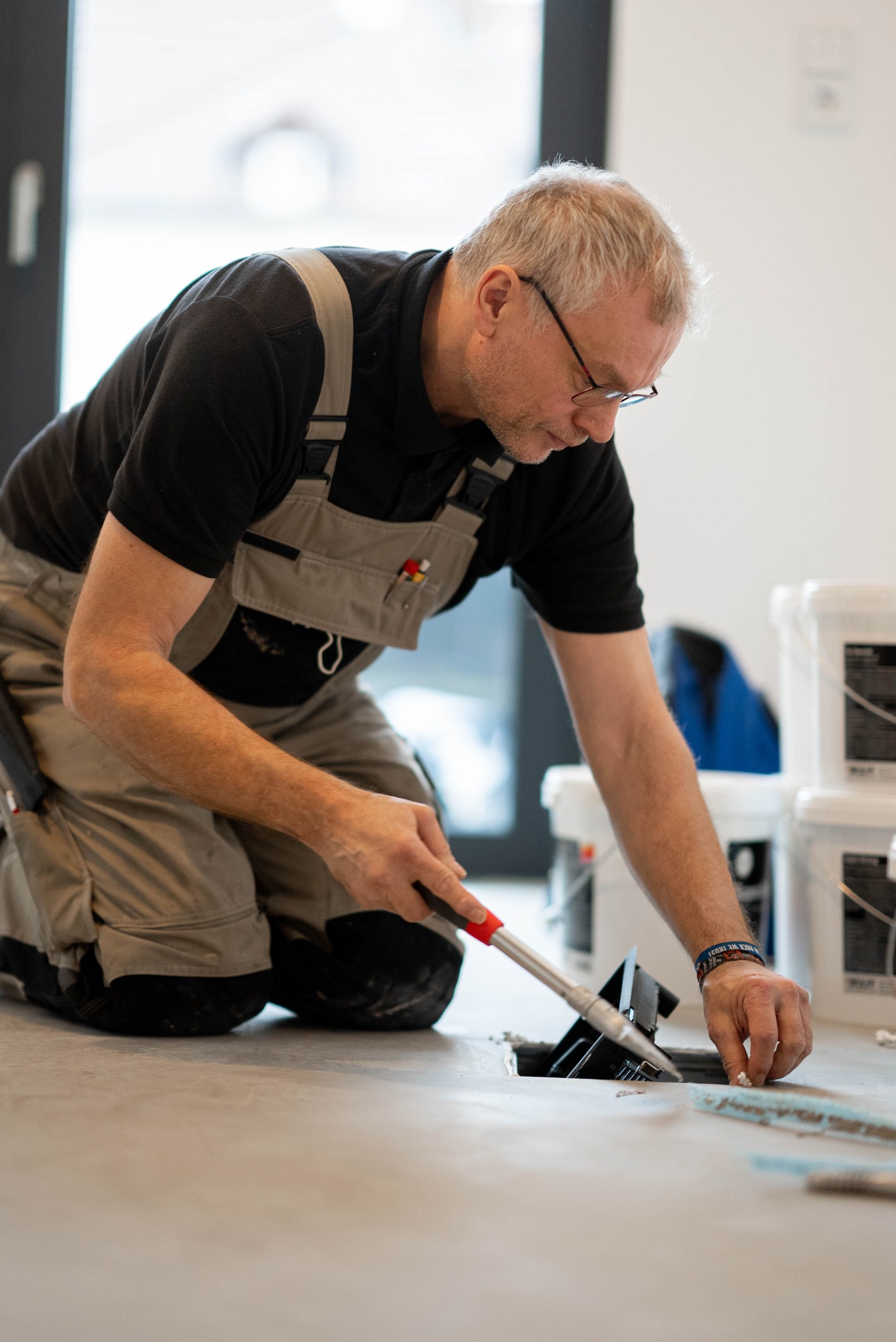 Ein Mann im Arbeitsoverall kniet auf dem Boden und trägt mit einer Kelle Klebstoff oder Fugenmasse auf, während er im Innenbereich Fußböden verlegt – ein wahrer Ausdruck unseres Engagements bei Über Uns für handwerkliches Können. - Farbenhaus Pflüger - Raumausstattung, Bodenbeläge, Vinylboden, Parkett, Farben, Malerarbeiten - Simmenet 2, 37235 Hessisch Lichtenau