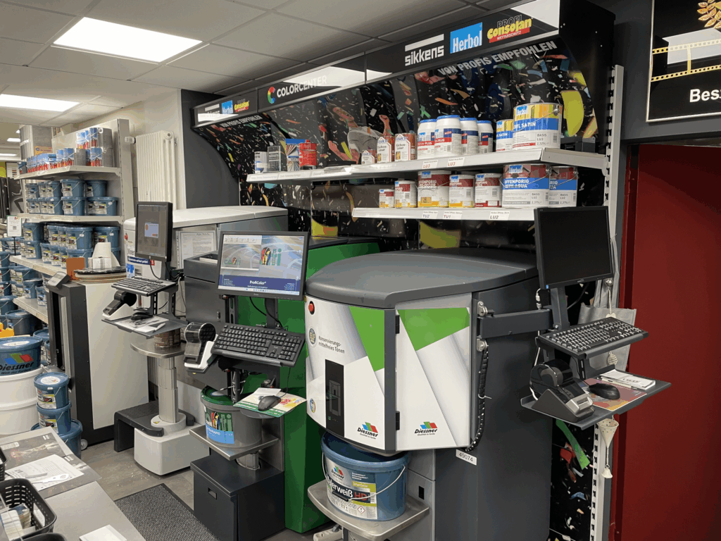 Farbmischstation mit Computern, Monitoren und verschiedenen Farbdosen und Zubehör auf Regalen im Inneren eines Ladens. - Farbenhaus Pflüger - Raumausstattung, Bodenbeläge, Vinylboden, Parkett, Farben, Malerarbeiten - Simmenet 2, 37235 Hessisch Lichtenau