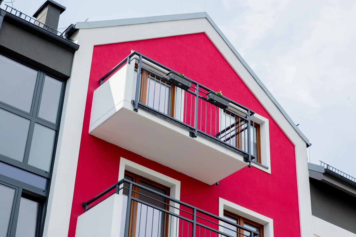 Ein modernes Gebäude mit leuchtend roter Fassade und zwei Balkonen mit schwarzem Geländer vor einem bewölkten Himmel. - Farbenhaus Pflüger - Raumausstattung, Bodenbeläge, Vinylboden, Parkett, Farben, Malerarbeiten - Simmenet 2, 37235 Hessisch Lichtenau