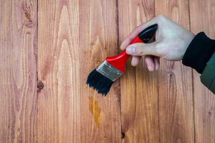 Eine Hand trägt mit einem Pinsel einen flüssigen Lack auf eine Holzoberfläche mit senkrechten Brettern auf. - Farbenhaus Pflüger - Raumausstattung, Bodenbeläge, Vinylboden, Parkett, Farben, Malerarbeiten - Simmenet 2, 37235 Hessisch Lichtenau