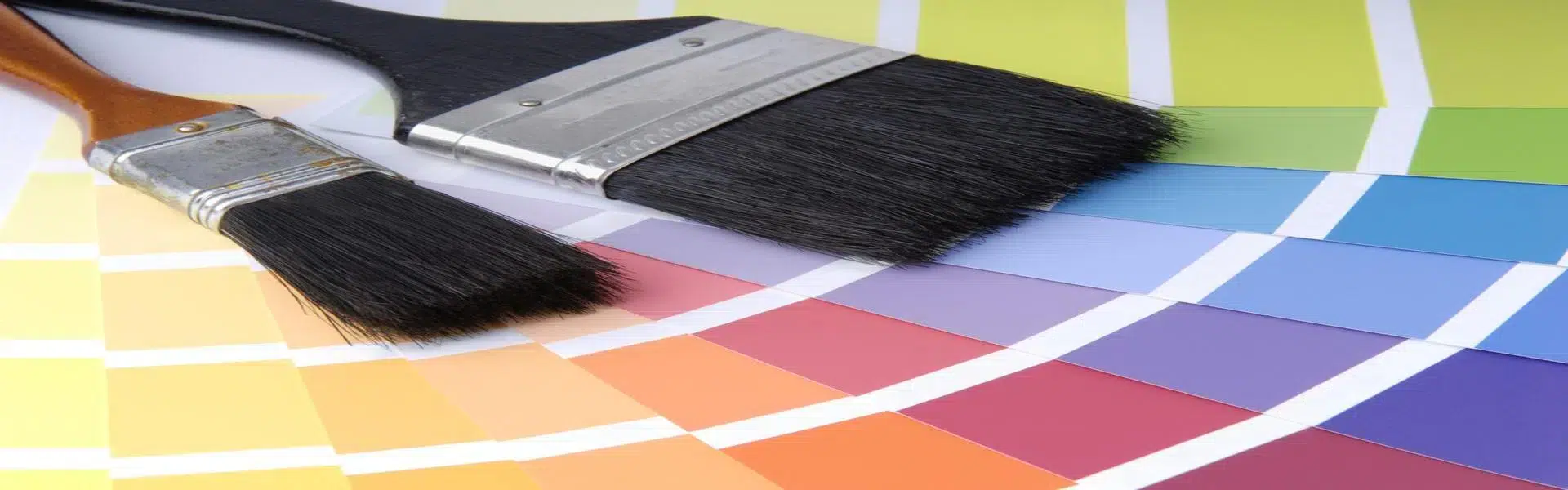 Zwei Pinsel liegen auf einem Spektrum bunter Farbmuster, die in einem fächerartigen Muster von Gelb über Rot und Lila bis hin zu Blau angeordnet sind. - Farbenhaus Pflüger - Raumausstattung, Bodenbeläge, Vinylboden, Parkett, Farben, Malerarbeiten - Simmenet 2, 37235 Hessisch Lichtenau
