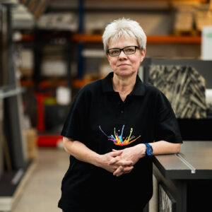 Eine Frau mit kurzen grauen Haaren und Brille steht in einem Arbeitsbereich. Sie trägt ein schwarzes Poloshirt mit einem farbenfrohen „Über Uns“-Design und eine blaue Uhr. Im Hintergrund sind Regale und Materialien zu sehen. - Farbenhaus Pflüger - Raumausstattung, Bodenbeläge, Vinylboden, Parkett, Farben, Malerarbeiten - Simmenet 2, 37235 Hessisch Lichtenau