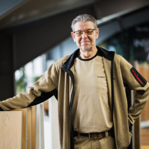 Gerhard Gallaus mit Brille und beiger Jacke steht im Innenbereich neben Holzpaneelen und blickt mit neutralem Gesichtsausdruck in die Kamera – perfekt für eine Über-Uns-Einführung. - Farbenhaus Pflüger - Raumausstattung, Bodenbeläge, Vinylboden, Parkett, Farben, Malerarbeiten - Simmenet 2, 37235 Hessisch Lichtenau