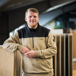 Kevin Hainke mit kurzen blonden Haaren und Bart steht drinnen, lehnt an einem Holzdisplay und trägt einen hellbraunen und schwarzen Pullover mit der Aufschrift „K. Halilaj“, und fängt mit seiner einladenden Präsenz den Geist von Über Uns ein. - Farbenhaus Pflüger - Raumausstattung, Bodenbeläge, Vinylboden, Parkett, Farben, Malerarbeiten - Simmenet 2, 37235 Hessisch Lichtenau