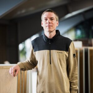 Dennis Knierim in einem beige-schwarzen Pullover steht drinnen, stützt eine Hand auf eine Holzoberfläche und blickt direkt in die Kamera – und fängt den zugänglichen Geist hinter unserer Über-Uns-Geschichte ein. - Farbenhaus Pflüger - Raumausstattung, Bodenbeläge, Vinylboden, Parkett, Farben, Malerarbeiten - Simmenet 2, 37235 Hessisch Lichtenau