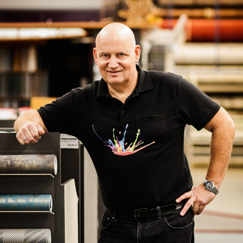 Ein Mann mit Glatze und einem schwarzen Hemd mit buntem Spritzermuster steht neben Stoffrollen in einer Werkstatt oder einem Laden von Über Uns. - Farbenhaus Pflüger - Raumausstattung, Bodenbeläge, Vinylboden, Parkett, Farben, Malerarbeiten - Simmenet 2, 37235 Hessisch Lichtenau