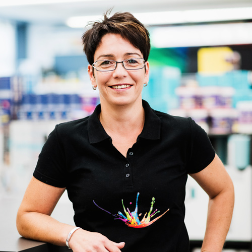 Eine Frau mit kurzen dunklen Haaren, Brille und einem schwarzen Hemd mit einem bunten Spritzermuster steht drinnen und lächelt in die Kamera – perfekt für unsere Über Uns-Einführung. - Farbenhaus Pflüger - Raumausstattung, Bodenbeläge, Vinylboden, Parkett, Farben, Malerarbeiten - Simmenet 2, 37235 Hessisch Lichtenau