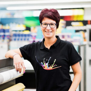 Melanie Kenn mit kurzen dunklen Haaren und Brille steht drinnen. Sie trägt ein schwarzes Hemd mit einem bunten Spritzermuster und steht neben einer großen Papierrolle – perfekt für den Abschnitt „Über uns“. - Farbenhaus Pflüger - Raumausstattung, Bodenbeläge, Vinylboden, Parkett, Farben, Malerarbeiten - Simmenet 2, 37235 Hessisch Lichtenau