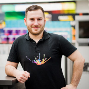 Ein Mann mit kurzen Haaren und Bart steht im Haus und lächelt. Er trägt ein schwarzes Poloshirt mit einem bunten Spritzer-Motiv. Einen Arm stützt er auf die Theke und verkörpert den freundlichen Geist unseres Über Uns-Teams. - Farbenhaus Pflüger - Raumausstattung, Bodenbeläge, Vinylboden, Parkett, Farben, Malerarbeiten - Simmenet 2, 37235 Hessisch Lichtenau