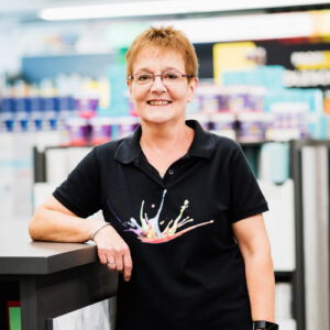 Eine Frau mit kurzen Haaren und Brille steht lächelnd an einer Theke im „Über Uns“-Bereich des Ladens und trägt ein schwarzes Poloshirt mit einem bunten Farbspritzer-Design. - Farbenhaus Pflüger - Raumausstattung, Bodenbeläge, Vinylboden, Parkett, Farben, Malerarbeiten - Simmenet 2, 37235 Hessisch Lichtenau