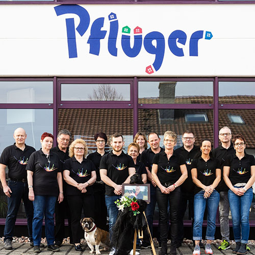 Team Farbenhaus Pflüger in passenden schwarzen Hemden steht vor einem Gebäude mit der Aufschrift "Pfluger", mit zwei Hunden und einem gerahmten Foto mit Blumen in der Mitte, versammelt für eine besondere Kontaktveranstaltung. - Farbenhaus Pflüger - Raumausstattung, Bodenbeläge, Vinylboden, Parkett, Farben, Malerarbeiten - Simmenet 2, 37235 Hessisch Lichtenau
